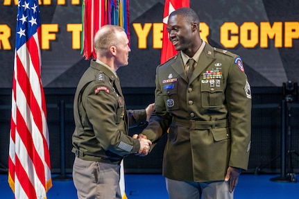An Army Soldier wearing green service dress uniform is shaking hands with another similarly dressed Soldier while standing in front of flags during a ceremony.