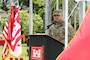 A male soldier stands at a podium. There is a group of flags on a stand in the background.