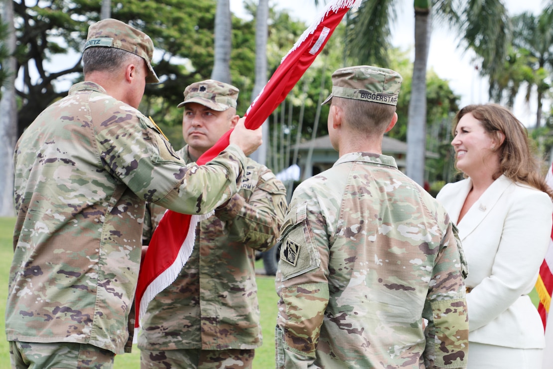 Three male Army Soldiers and one female stand facing each other. One is handing a red flag wrapped around a pole to another. There are palm trees in the background