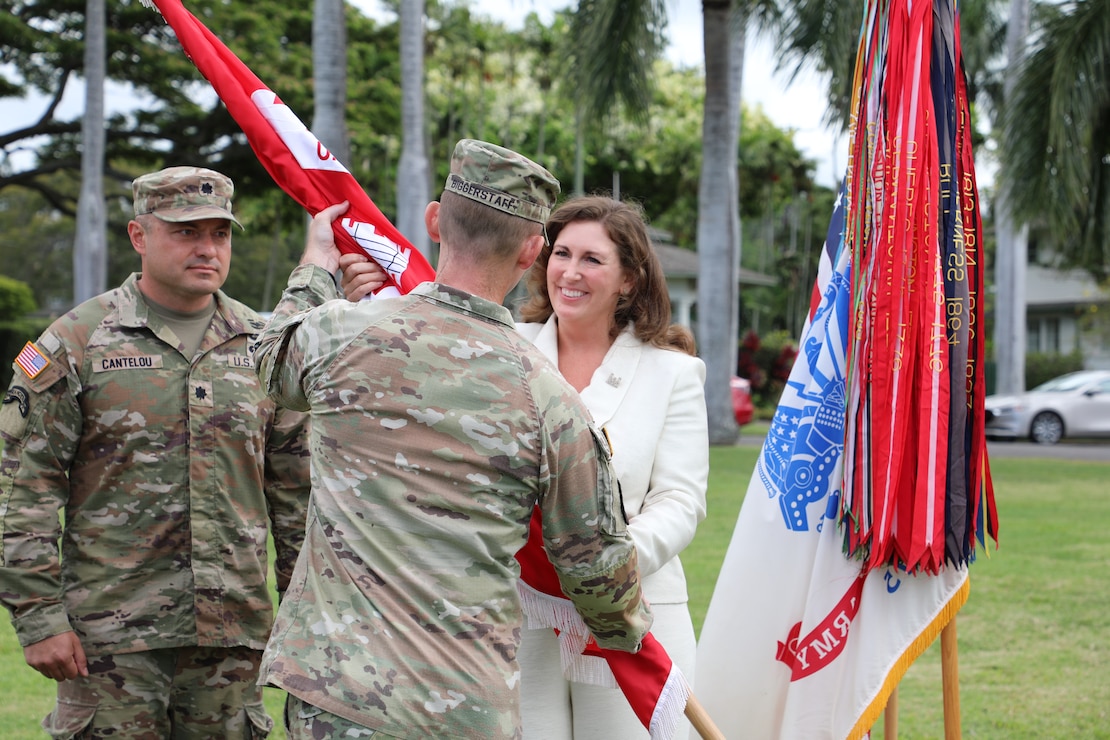 Back of a male Army Soldier is grasping a red flag wrapped around a pole from a smiling female in an off-white pants suit.