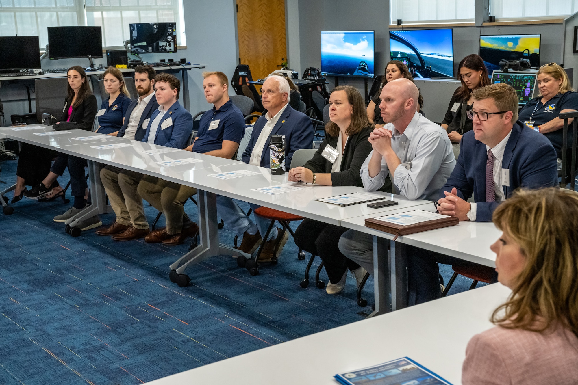 A group of men and women, wearing business casual attire, sit along a long table. Large LCD monitors can be seen in the background