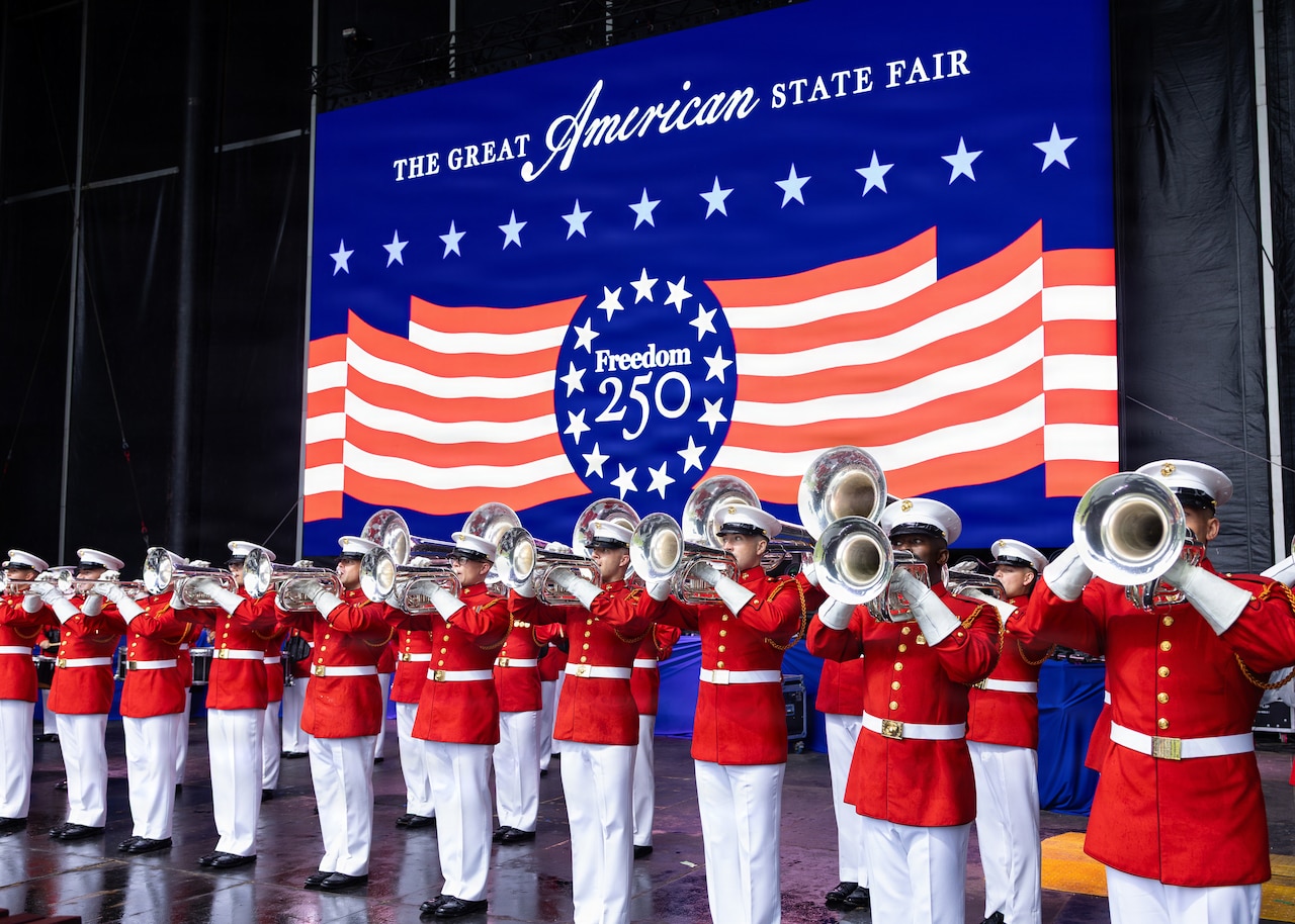 Military musicians wearing Marine dress uniforms play instruments. A large banner behind them reads "The Great American State Fair."