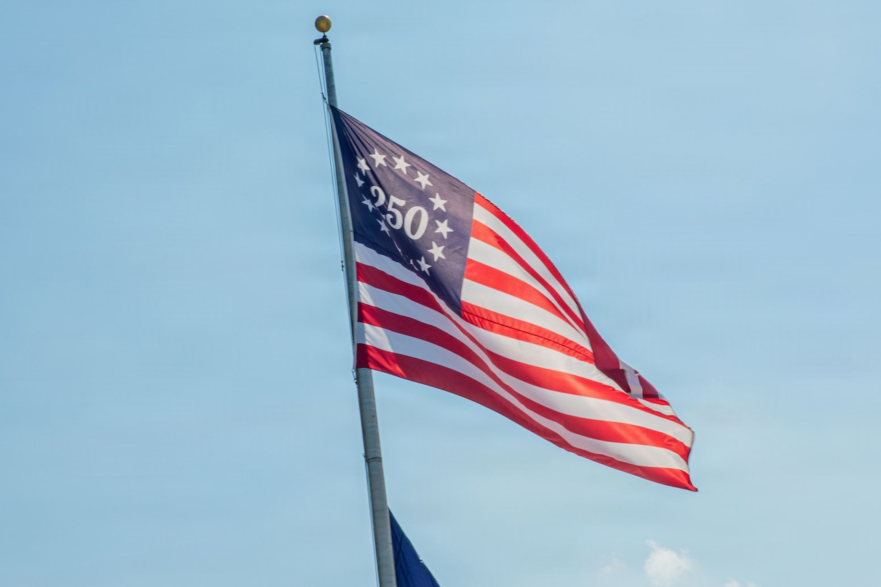 A commemorative flag flies against the backdrop of a blue sky. The flag has 13 stars and the number "250" where there would normally be 50 stars on an American flag.