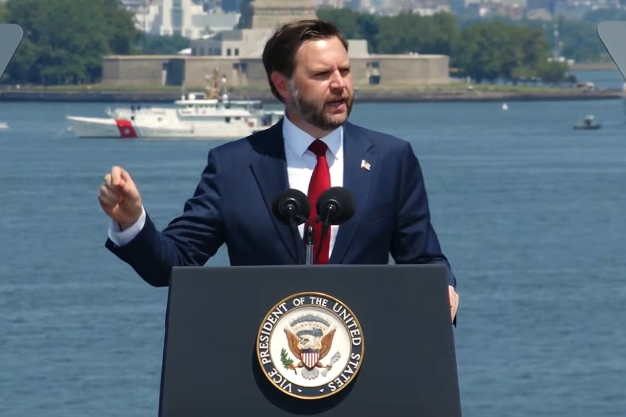 A man stands behind a lectern. In the background are ships in a harbor, high-rise buildings and the base of the Statue of Liberty.