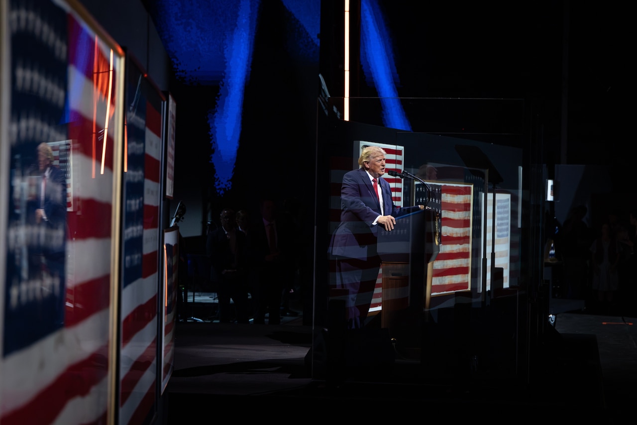 A man wearing a business suit stands behind a lectern and speaks. Behind him are various versions of the American flag.