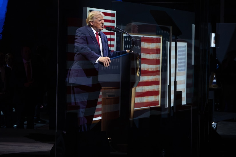 A man wearing a business suit stands behind a lectern and speaks. Behind him are various versions of the American flag.