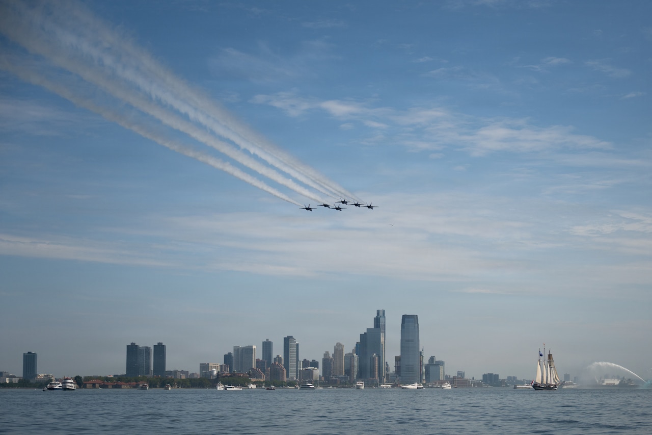 Several aircraft fly over a river and city skyline in formation with several vessels in the water.