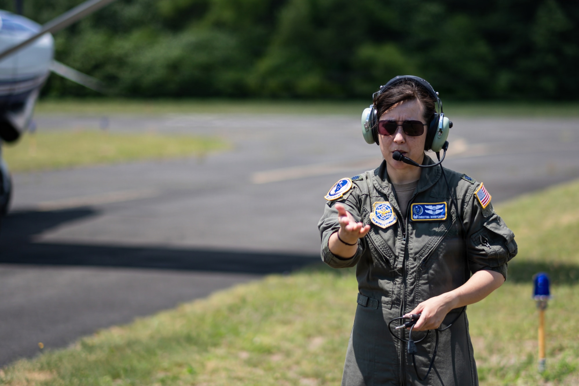 A woman in a flight duty uniform and sunglasses gestures in front of a small aircraft.