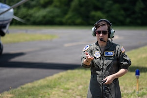 A woman in a flight duty uniform and sunglasses gestures in front of a small aircraft.