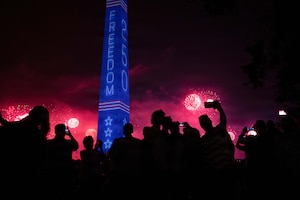 Spectators in silhouette in the foreground watch red fireworks in the sky near a tall monument covered in blue with white stars.