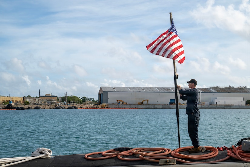 A man wearing a dark blue military uniform raises an American flag while standing on the deck of a submarine; water and buildings are in the distance.
