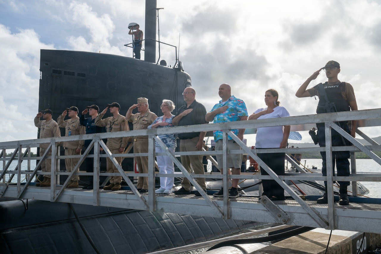 Nearly a dozen people wearing military and civilian attire salute while standing on the gangway of a submarine.