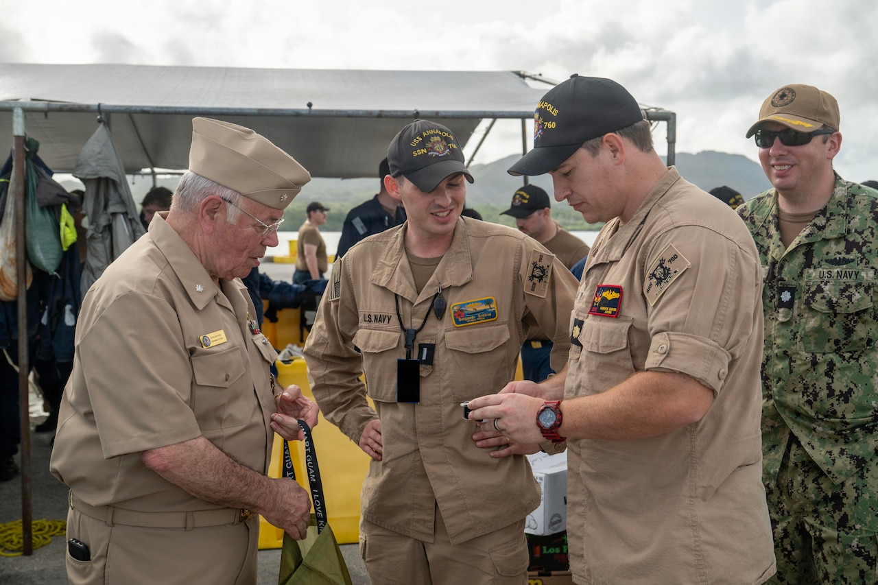 A man in a formal military uniform presents another man in similar attire with a gift while two others smile and watch.