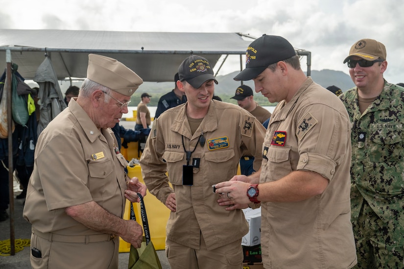 A man in a formal military uniform presents another man in similar attire with a gift while two others smile and watch.