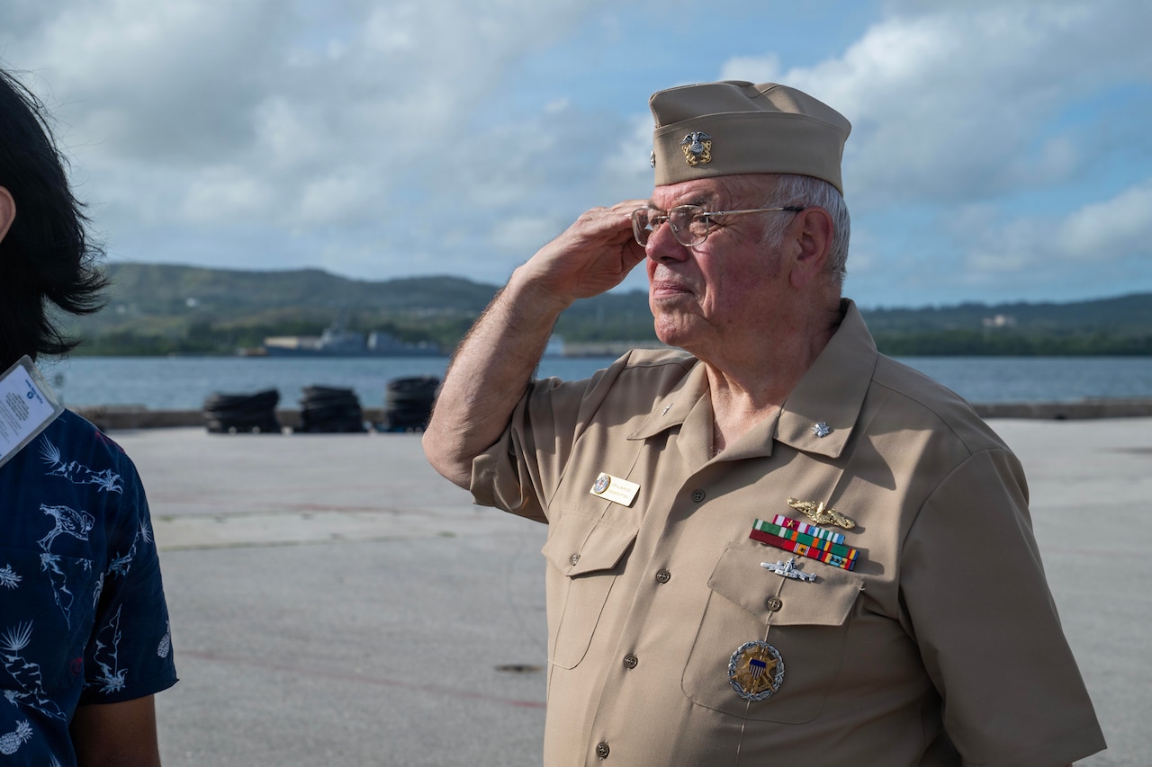 A man in a formal military uniform salutes an off-camera flag; mountains and water are in the distance.