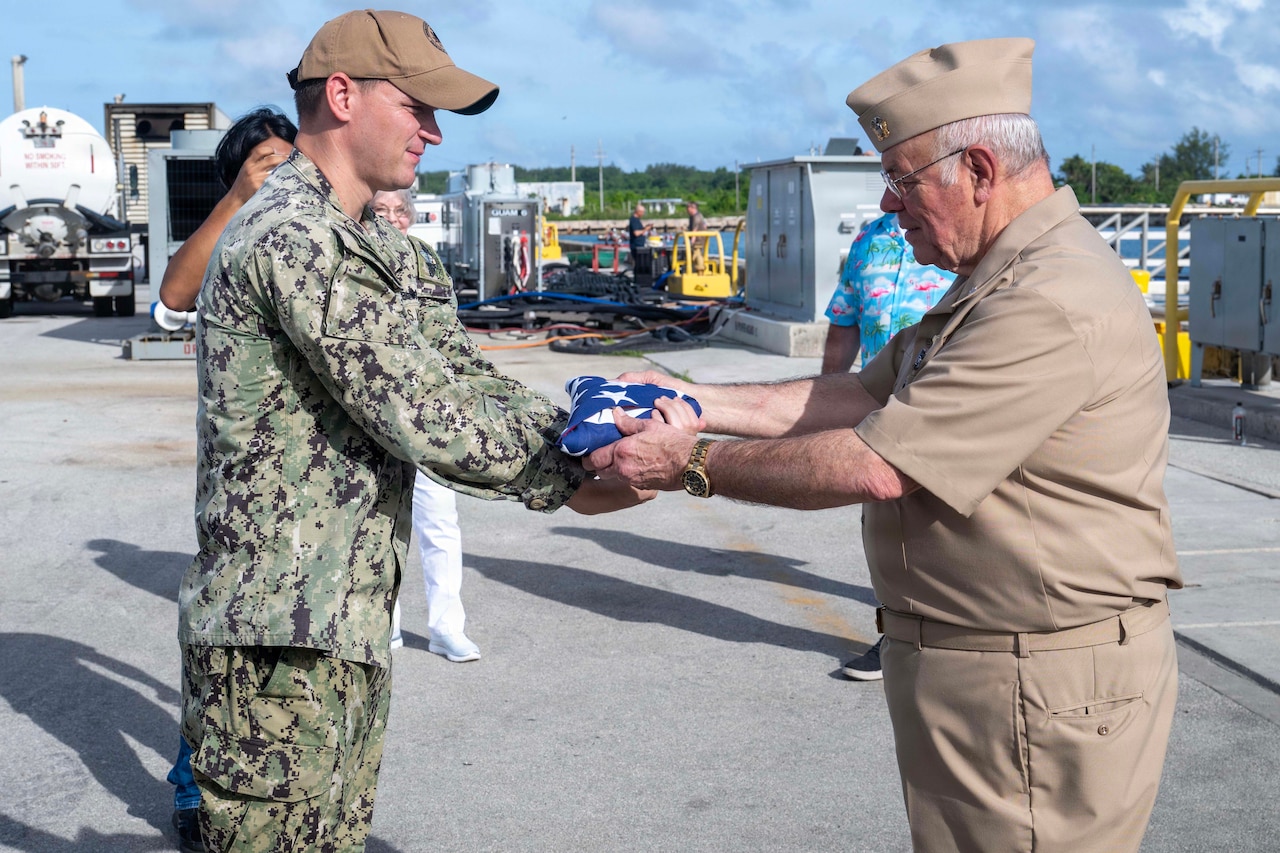 A man in a camouflage military uniform presents a folded American flag to a man in a formal military uniform; heavy equipment and transport containers are in the background.