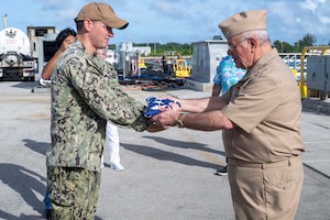 A man in a camouflage military uniform presents a folded American flag to a man in a formal military uniform; heavy equipment and transport containers are in the background.