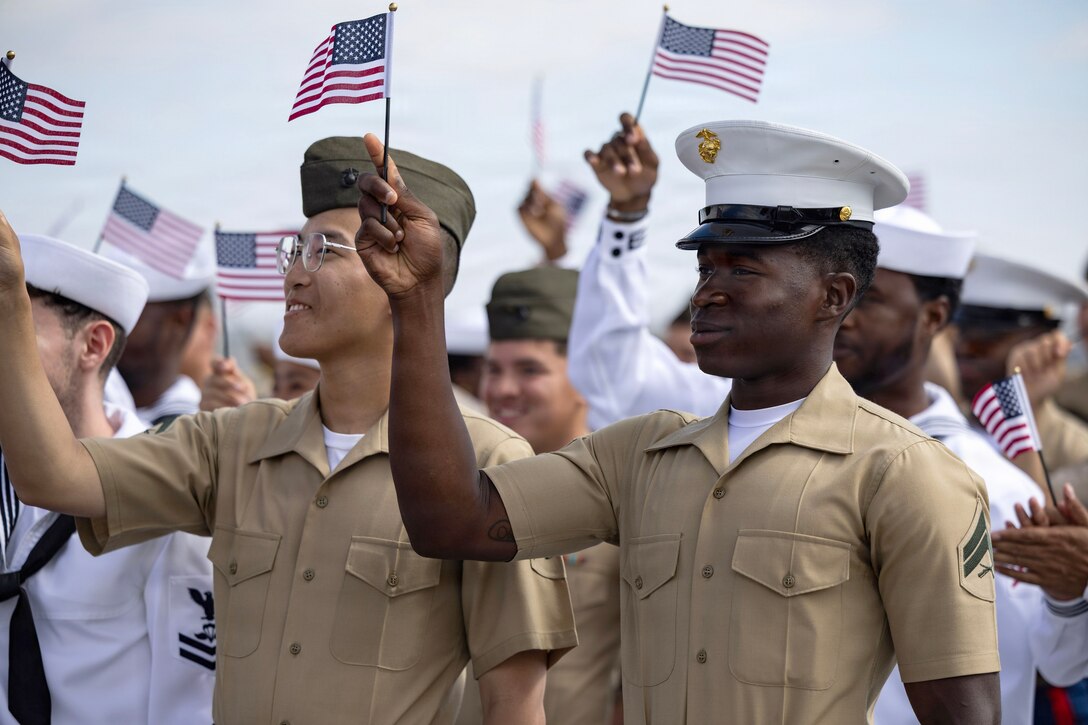 A large group of Marines and sailors wave American flags against a blue sky.
