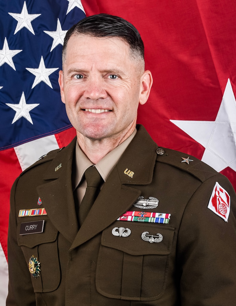 A man in an Army brigadier general uniform poses for a photo in front of the U.S. and brigadier general one-star flags