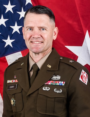 A man in an Army brigadier general uniform poses for a photo in front of the U.S. and brigadier general one-star flags