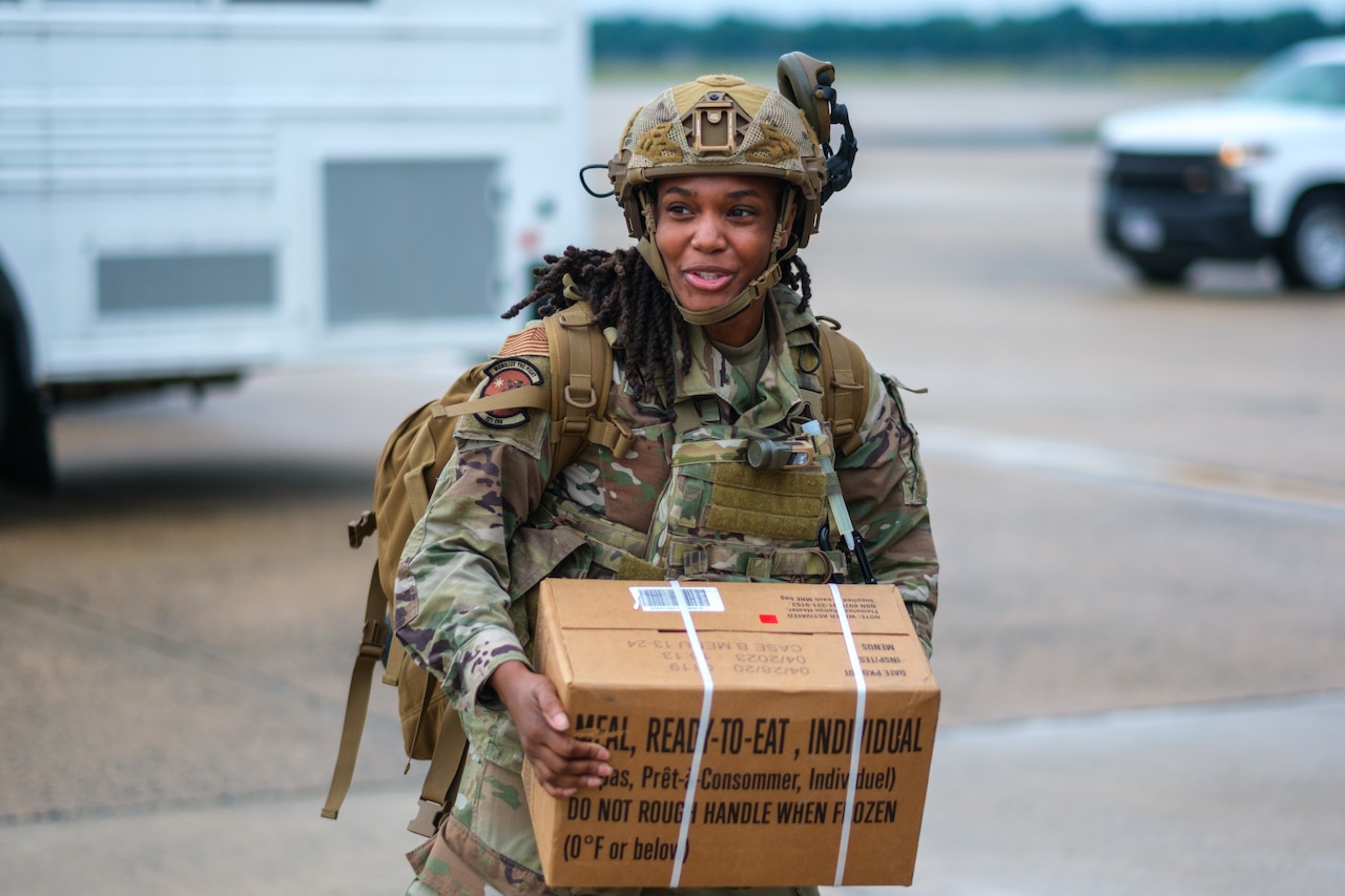 An airman wearing camouflage uniform and helmet carries a case of military rations with vehicles parked in the background.
