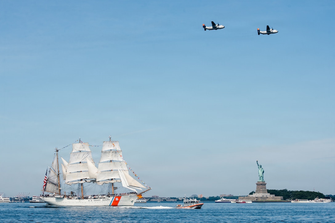 A three-masted sailing ship with white sails passes the Statue of Liberty as two propeller-driven military aircraft fly overhead.