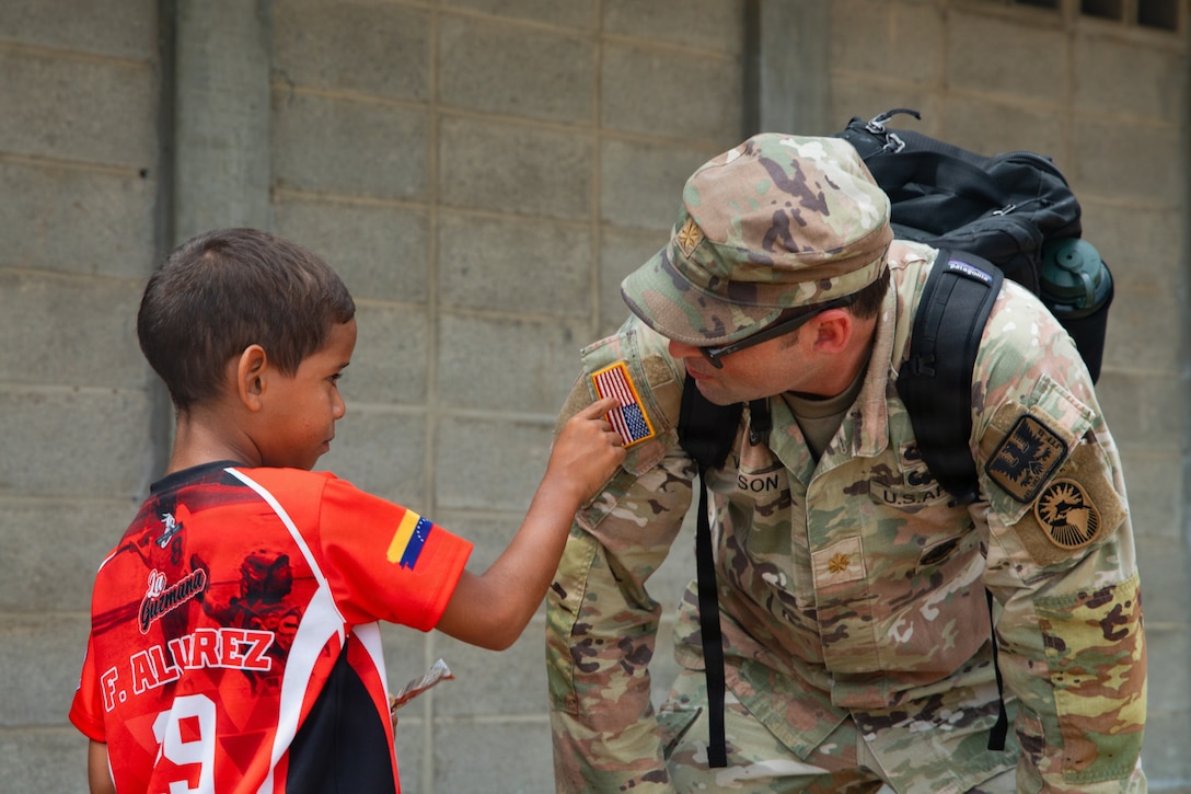 A boy points to the American flag on the shoulder of a soldier who is bending over to speak to him.