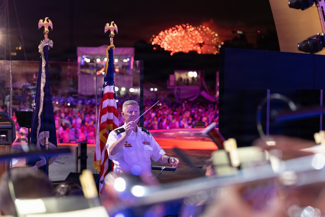 A conductor waves his baton in front of an orchestra with a large crowd and fireworks in the background of an outdoor setting.