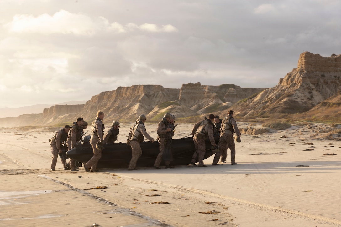 A group of about a dozen Marines in camouflage uniforms drag a rubber boat onto a deserted beach with rugged hills in the background.