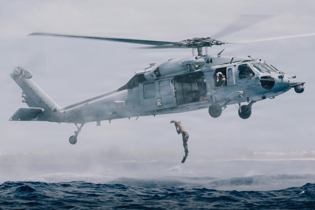 A sailor wearing fins jumps feet first into a body of choppy water from the open doorway of a hovering helicopter during daytime.