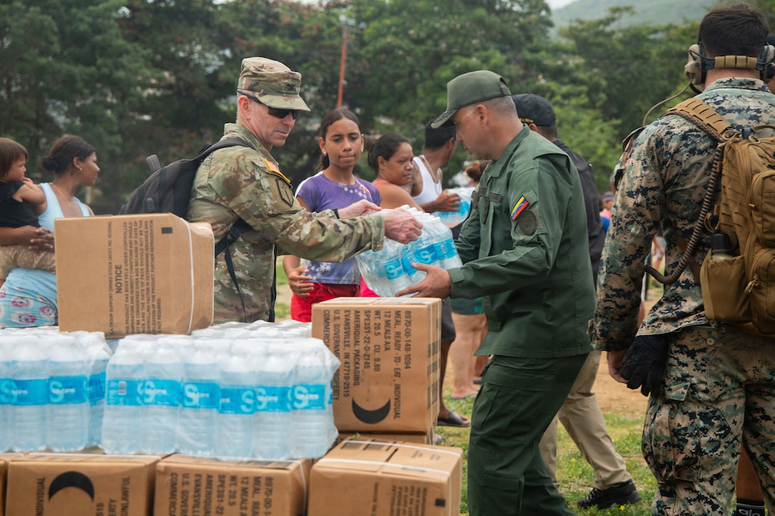 A U.S. soldier in a camouflage uniform hands a plastic-wrapped case of water bottles to a Venezuelan service member as people in civilian attire in the background wait to receive it outdoors during daytime; there are boxes and more water bottles stacked in the foreground.