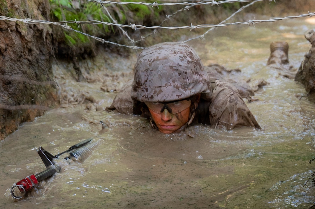 A Marine in combat gear crawls in a watery trench with barbed wire strewn across it during daytime.