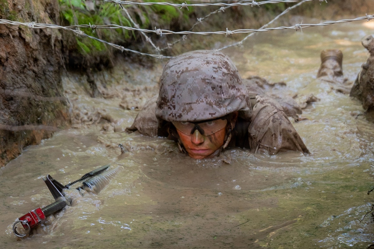 A Marine in combat gear crawls in a watery trench with barbed wire strewn across it during daytime.