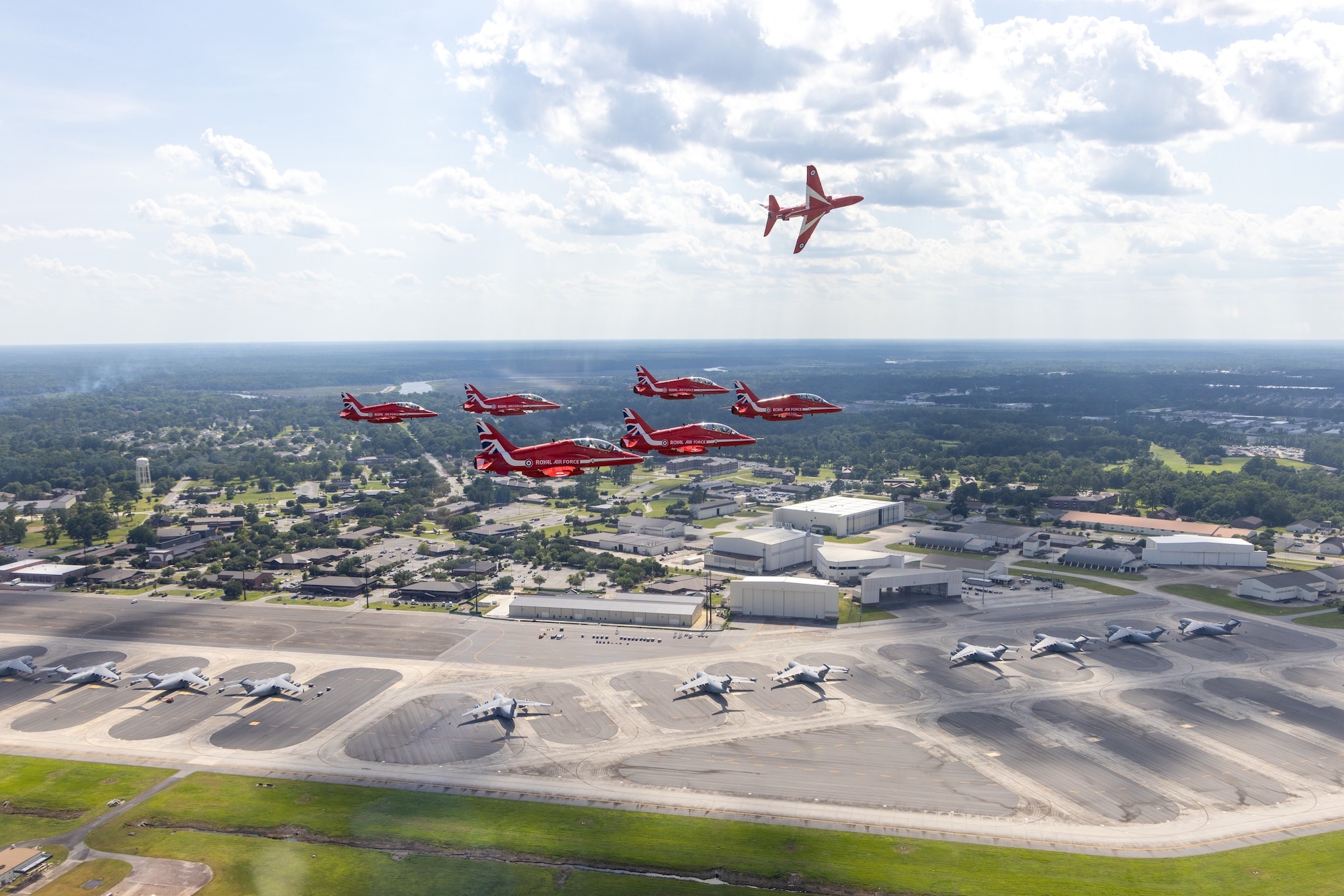 Six planes fly in a triangle formation, one plane flies on its side above the formation. Below them is a flightline with parked C-17s, and the base with buildings and trees.
