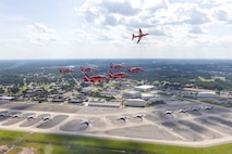 Six planes fly in a triangle formation, one plane flies on its side above the formation. Below them is a flightline with parked C-17s, and the base with buildings and trees.