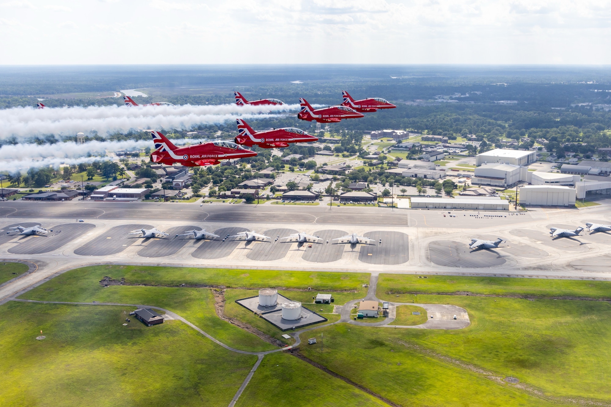 Six planes fly in a triangle formation with condensation trails behind them. Below them is a flightline with parked C-17s, and the base with buildings and trees.