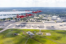Six planes fly in a triangle formation with condensation trails behind them. Below them is a flightline with parked C-17s, and the base with buildings and trees.