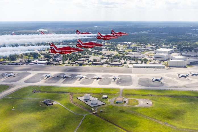 Six planes fly in a triangle formation with condensation trails behind them. Below them is a flightline with parked C-17s, and the base with buildings and trees.