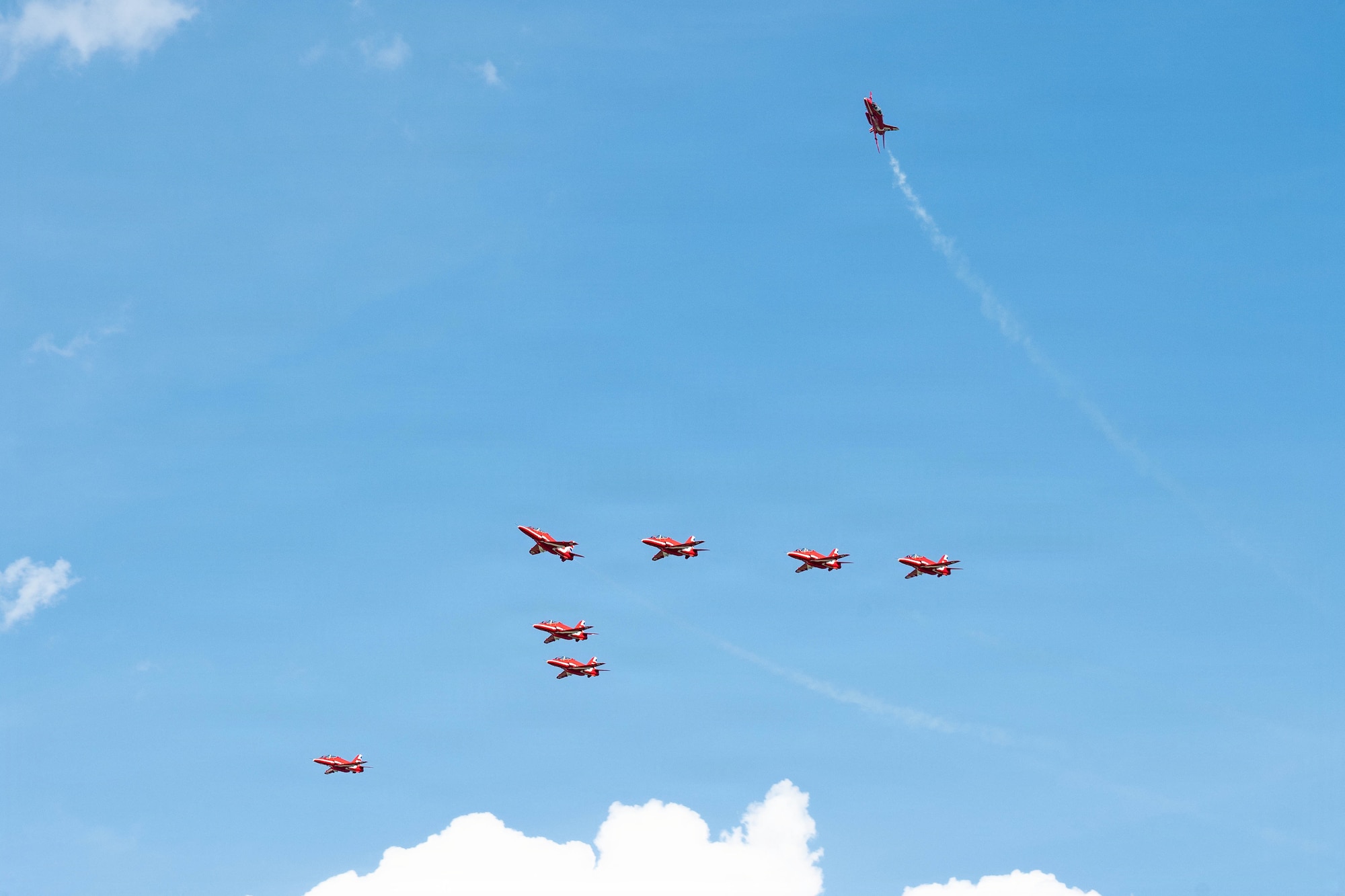 Six red planes fly in a triangle formation, one red plane curves away out of formation and one red plane flies next to the formation.