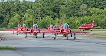 Five red planes drive in two columns, two turn in to join the lines behind them. Trees are visible in the background.