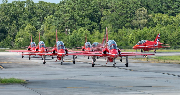 Five red planes drive in two columns, two turn in to join the lines behind them. Trees are visible in the background.