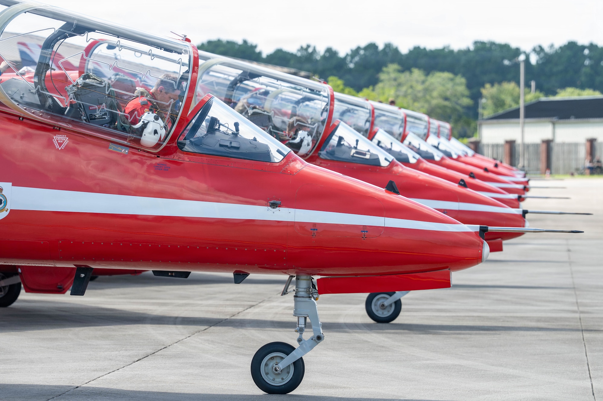 Seven red planes sit in a row, the tops are open and a building and trees are visible in the background.
