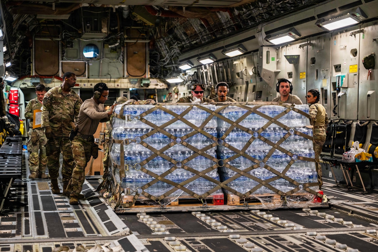 Several airmen push a large pallet of supplies from inside the back of a large cargo aircraft.