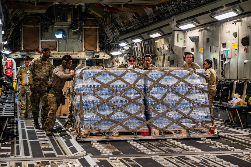 Several airmen push a large pallet of supplies from inside the back of a large cargo aircraft.