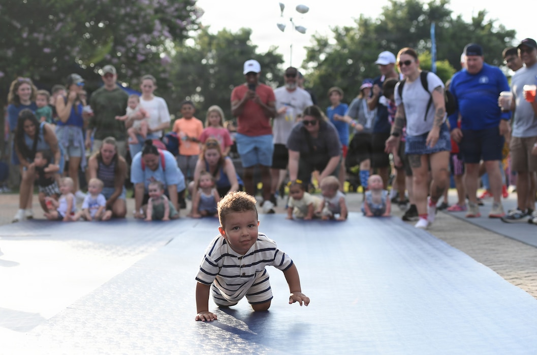 A baby crawls on a mat in front of a group of people.