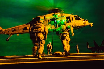 Two sailors wearing tactical gear and helmets with green lights run toward a helicopter hovering over the deck of a ship in dark conditions, as another sailor in similar attire stands near a rope attached to the helicopter, with a hazy, green sky in the background.