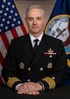 Caucasian male wearing navy dress uniform is posed with hands on top of each other and sitting in front of a US and Navy flag.