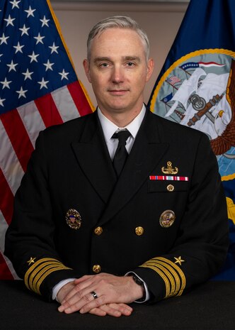 Caucasian male wearing navy dress uniform is posed with hands on top of each other and sitting in front of a US and Navy flag.