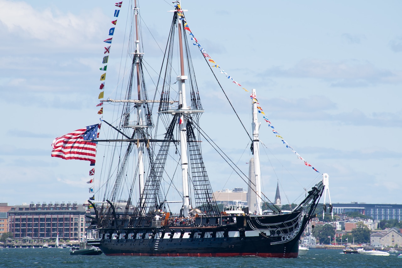 A large 18th-century ship sails in a harbor on a cloudy day while flying the American flag; there are buildings in the background.