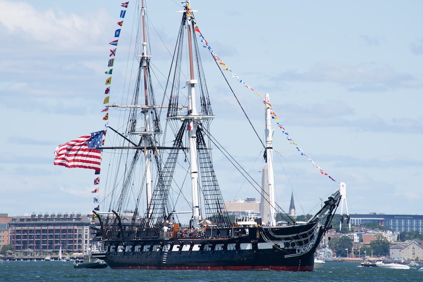 A large 18th-century ship sails in a harbor on a cloudy day while flying the American flag; there are buildings in the background.
